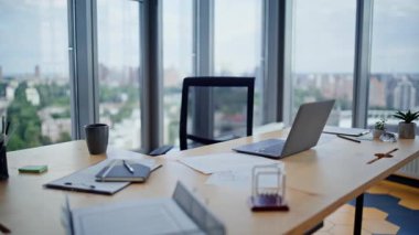 Şehir manzaralı modern ofiste dizüstü bilgisayarı olan boş bir şirket çalışma alanı. Profesyonel ahşap masaya yerleştirilmiş kahve fincanı. Üretken iş akışı için panoramik pencereli işyeri içi.