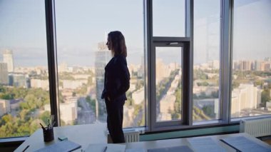 Ofis panoramik penceresinden şehir manzarasına bakan bir işletmeci. Cam duvarın önünde duran zarif bir iş kadını. Çalışmayı düşünen yüksek binalar. Kendine güvenen kadın CEO çalışma alanından şehir manzarasını seyrediyor..