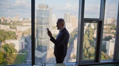 Ofis panoramik penceresinin yanında akıllı telefonlara bakan aşırı neşeli iş adamı. Heyecanlı yaşlı yönetici harika bir cep telefonu mesajı iletiyor. Duygusal çalışan el kaldırıyor. Telefonu tutmada başarılı oluyor.