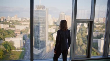 Modern ofiste panoramik şehir manzaralı şirket çalışanı. Yüksek binadan şehir manzarası seyreden ciddi bir iş kadını. Seçkin takım elbiseli kadın CEO koridorda tek başına dinleniyor..