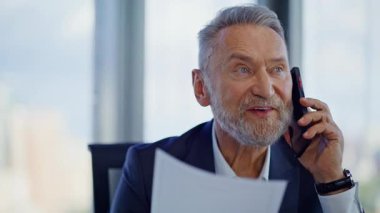 İş adamı cep telefonuyla belgeyi tartışırken ofis kapanışında başarılı bir anlaşmanın keyfini çıkar. Gülümseyen yaşlı müdür elinde kağıtlarla cep telefonundan konuşuyor. Neşeli yönetici akıllı telefondan harika haberler paylaşıyor..