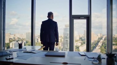 İşyeri sahibi modern ofis işyerinden şehir manzarası düşünüyor. Kendine güvenen kıdemli işadamı panoramik pencerenin yanında şehir manzarasına bakıyor. Şık takım elbise tasarlamada başarılı bir lider..