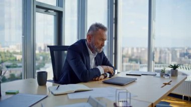 Yaşlı girişimci stresli bir şekilde modern işyerinde kapalı bir dizüstü bilgisayarla oturuyor. Finansal kriz üzerine düşünen sakallı bir işadamı çözüm arıyor. Endişeli yaşlı yönetici iş sorunları yaşıyor