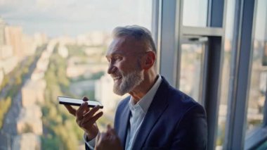 Hoparlörden konuşan yaşlı girişimci panoramik pencerenin önünde bekliyor. Gülümseyen işadamı hoparlörden arıyor. Mutlu kıdemli yönetici sesli mesaj kaydediyor.