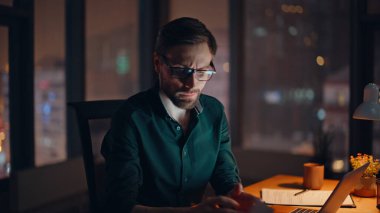 Panoramik pencere kenti manzaralı gece ofisinde çalışan profesyonel analist. Sakallı işadamı lamba ışığında dizüstü bilgisayarına bakıyor. Finansal raporu analiz eden ciddi yatırımcı.