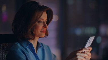 Fazla mesai yöneticisi karanlık iş yerinde SMS cep telefonunu okuyor. Güzel bir iş kadını gece ofisinde online akıllı telefon uygulamasına göz atıyor. Gülümseyen kadın çalışan gece geç saatlerde cep telefonuna mesaj atıyor..