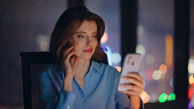 Karanlık ofisteki akıllı telefon mesajlarını okuyan geç çalışan bir çalışan. Şehir ışıkları manzaralı. Gülümseyen iş kadını gece internet cep telefonu uygulamasına bakıyor. İş yerinde sosyal medya izleyen rahat bir kadın..