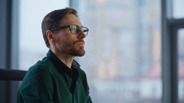 Başarılı yönetici, modern kurumsal iş yerini kamerayla işaret ediyor. Ofis penceresinde poz veren gülümseyen iş adamı portresi. Gözlüklü kendine güvenen yönetici çalışma yerinde oturuyor.