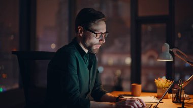 Gece iş yerinde gülümseyen serbest çalışan biri. Yakın plan. Mutlu sakallı iş adamı akıllı telefon konuşmasına bilgisayarla karanlıkta oturarak başlar. Gözlüklü adam geç saatte cep telefonuyla konuşuyor..