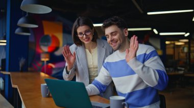 Gülümseyen takım arkadaşları ofis alanında dizüstü bilgisayarla video görüşmelerini selamlıyorlar. Şirket işyerindeki online ekip toplantısında olumlu kadın konuşmaları. Üretken uzaktan beyin fırtınası sağlayan iş arkadaşları 