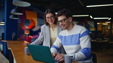 Gülümseyen iş arkadaşları modern kafeterya tezgahında dizüstü bilgisayarı görüntülüyorlar. Profesyonel kadın, proje detaylarını müşteriye anlatıyor. Neşeli takım çalışması içeride sanal toplantı hakkında konuşuyor.