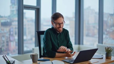 Gülümseyen yönetici video konferansında danışmanlık yapıyor. Modern ofiste laptop web kamerasına kağıtları gösteriyor. İnternetten konuşan gözlüklü profesyonel bir iş adamı. Mutlu çalışanın sanal toplantısı var.