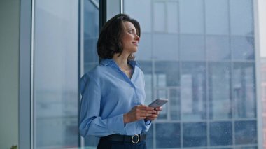 Ofis penceresinin yanındaki cep telefonu mesajını okuyan mutlu bir çalışan. Gülümseyen kadın yönetici akıllı telefondan maaş bildirimi alıyor. Çevrimiçi telefon uygulamasına göz atan zarif bir iş kadını.