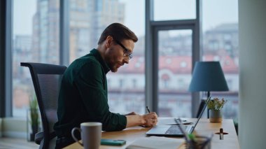Işık ofisindeki iş projesinde dizüstü bilgisayarla çalışan yaratıcı girişimci makaleleri. Gülümseyen yetenekli analist, masadaki notları kontrol ediyor. Başarılı iş adamı açılışı analiz ediyor.