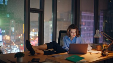 Gece çalışma alanında lamba ışığında başarılı bir bayan dizüstü bilgisayara göz atıyor. Ofis panoramik penceresinin yanında oturan parlak şehir manzaralı gülümseyen bir iş kadını. Mutlu kadın CEO bacakları masaya koyuyor..