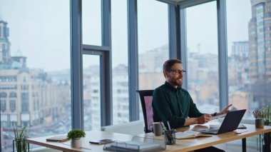 Kendine güvenen yönetici, modern kurumsal işyerinin dizüstü bilgisayarında başarılı olduğunu düşünüyor. Finans raporunu okuyan gülümseyen iş adamı gözlüğünü çıkarıyor. Ofis panoramik penceresinde çalışan işletme yöneticisi.