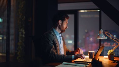 Gece apartmanında dizüstü bilgisayarda çalışan odaklanmış bir adam. Ciddi bir adam akşam evinde bilgisayar izlerken notları kontrol ediyor. Çevrimiçi yönetici e-postaya uzaktan cevap veriyor. Uzaklık iş konsepti 