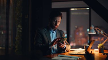 Koyu renk daire kapanışında cep telefonu uyarılarını kontrol eden serbest çalışan bir adam. Ciddi sakallı iş adamı akıllı telefon mesajı okuyor ve gece ofisinde notlar yazıyor. Geç saatte yönetici yalnız çalışıyor.