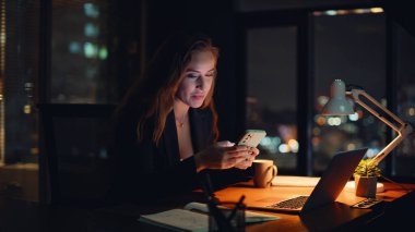 Kadın serbest çalışan cep telefonuna mesaj atıp karanlık bir yerde oturuyor. Gülümseyen iş kadını gece ofisinde akıllı telefon mesajı okuyor. Rahatlamış yönetici cep telefonunda sosyal medyaya göz atıyor