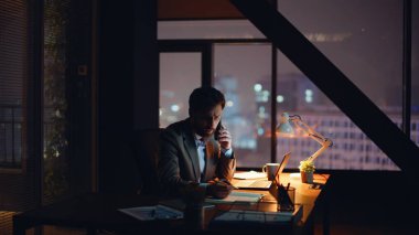 İdari vardiya kabinesini arıyorum. Endişeli adam, panoramik karanlık iş yerinde cep telefonundan konuşuyor. Stresli iş danışmanı elinde dizüstü bilgisayarla ofis masasında oturuyor.