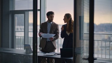 Belgeleri inceleyen iş adamları modern mimari bürosunda sohbet ediyorlar. Ciddi ortaklar panoramik pencere içinde konuşuyor. Neşeli adam kadın meslektaşına kontrat belgelerini gösteriyor.