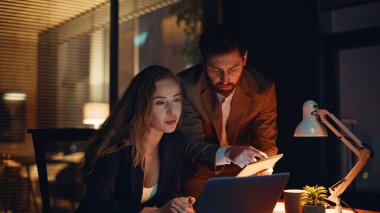 Birlikte iş yaptığı için mutlu iş arkadaşları. Gülümseyen kadın, akşam ofisinde bilgisayarla başarılı çalışma konusunu tartışıyor. Tableti kontrol eden iki iş arkadaşı kabinede ortaklığın keyfini çıkarıyor 