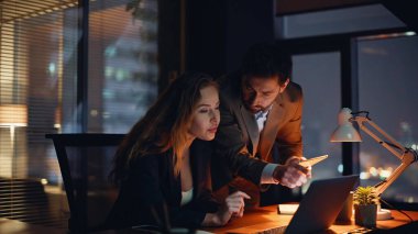 Fazla mesai başlatanlar akşam çalışma alanında strateji tartışıyorlar. Birlikte dizüstü bilgisayarı yakından izliyorlar. Profesyonel yöneticiler gece proje planlıyorlar. İki iş arkadaşı iş birliği yapıyor. 