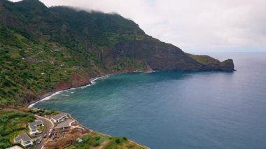Rocky coastline meeting ocean water drone shot. Breathtaking sea waves washing rough coastal mountain slopes covered lush greenery. Small settlement standing on green highland. Calm marine vacation.