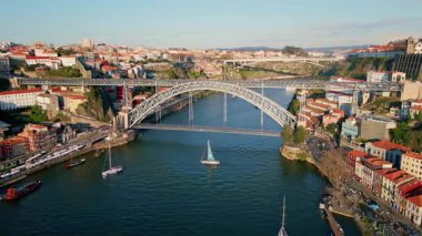Drone view steel bridge in Porto stretching over Douro river. Historic monument connecting tiled rooftops on shores. Boats navigating under metal arch on calm water surface. Dom Luis landmark Portugal