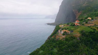 Gloomy marine landscape drone view. Tiled roof rural buildings standing on high coastal rocks cloudy day. Small settlement placed on green mountain hills raising over calm ocean water. Seascape beauty