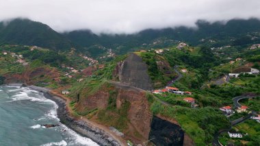 Breathtaking rocky shore drone view. Lush greenery covering mountain hills washed by beautiful ocean waves. Blue sea water splashing foaming on dark stone beach. Picturesque cloudy coastal landscape.