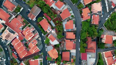 Red tiled houses cluster along narrow road network. Drone view neighborhood structure with green vegetations. Cozy village building with bright rooftops standing in rows. Residential architecture.