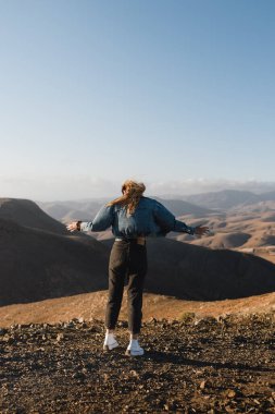 Genç bir kadın kendini özgür hissediyor rüzgarda uzun saçlarıyla ve şehir kıyafetiyle Fuerteventura 'daki bir çöl tepesinde günbatımının tadını çıkarıyor.