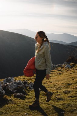 Uzun saçlı, güneş gözlüklü ve dağ elbiseli bir kız gün batımında gülümseyerek dağda yürür..