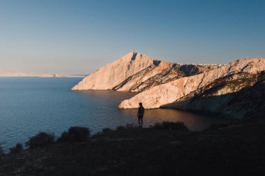 Yunan folegandros adasının kayalıklarının panoramik manzarası bir adamın silüetiyle gün batımının ılık renkleriyle aydınlandı