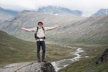 Happy hiker celebrating on a rock with arms open in a scenic Norwegian mountain valley. clipart