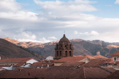 Cusco şehir manzarasında kilise kulesi ve terracotta çatıları görünüyor. Arka planda bulutlu gökyüzünün altında dağlar var.