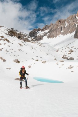 A hiker wearing snowshoes treks across a snowy landscape in the Picos de Europa. A vibrant turquoise glacial pond adds a pop of color against the pristine white snow and rugged mountain backdrop, creating a breathtaking scene of winter exploration. clipart