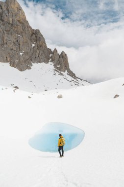 Yalnız bir maceracı, Picos de Europa 'da el değmemiş kar ve yüksek uçurumlarla çevrili canlı mavi bir buzul göletinin yanında duruyor. Sahne, dağlarda kışın dingin güzelliğini yakalar..