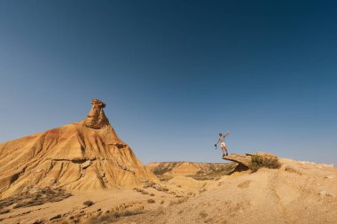 Bir gezgin kayalık bir uçurumun kenarında duruyor, kolları geniş, Bardenas Reales 'in nefes kesen çöl oluşumlarına tepeden bakan canlı mavi gökyüzü altında..