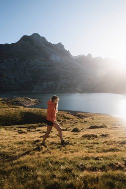 Güneşin doğuşunda Pireneler 'de bir dağ gölünün yanında yürüyen kadın yürüyüşçü, nefes kesici manzaranın tadını çıkarıyor.