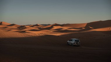 Arazi aracı, Merzouga Çölü, Fas 'ta batan güneşin sıcacık parıltısıyla yıkanmış, dalgalı erg chebbi kum tepeleri arasında duruyor.