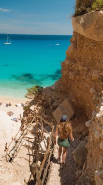 Woman hiking down rocky path to cala goloritze's turquoise waters clipart