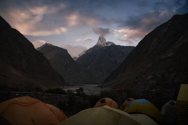 Baltoro Vadisi Karakoram Karakoram 'da, gün batımında dramatik bir gökyüzünün altında görkemli karlı bir dağ zirvesine bakan renkli kamp çadırları bir vadide duruyor.