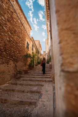 Bir gezgin, açık mavi gökyüzünün altında, kırsal binalar ve canlı saksılarla çevrili Fornalutx, Mallorca 'nın büyüleyici taş basamaklarından yükselir..