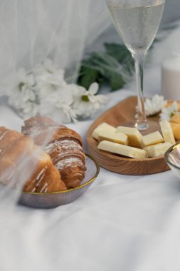 Çıtır kruvasanlar cam bir kasede beyaz çikolata, şarap ve çiçeklerin yanında beyaz kumaşın arka planında yatıyor. Romantik randevu pikniği, dikey yakın çekim