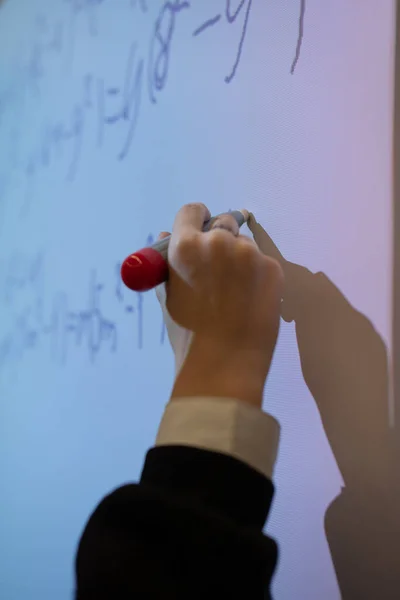 The hand holds a gray stylus with a red tip and writes a mathematical formula for a quadratic equation on a multimedia board during an algebra lesson. Close-up, vertical frame