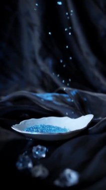 Mavi boncuklar arka planda parlayan siyah kumaşın üzerine yavaşça düşer, beyaz küçük dekoratif bir plakaya yansıyarak sihirli bir şekilde etrafa, dikey çekim yapar.