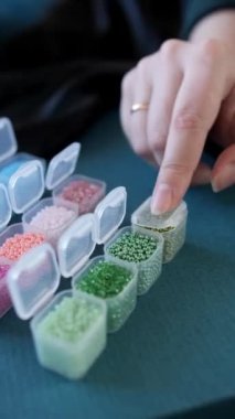 Parmaklarıyla boncuklu dokuma ustası bir kadının eli sırayla farklı renklerde boncuklarla dolu ve kapaklarla birbirine bağlı küçük plastik kapları kapatır. Yakın çekim, dikey çerçeve