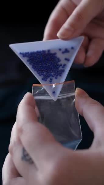 女性のビーズ織人の手は青いビーズと三角形の容器を保持し それを注ぎ 小さな透明なビニール袋に詰め それを閉じます 縦型クローズアップ撮影 — ストック動画